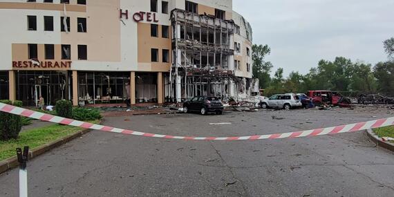 A building bearing the words "hotel" and "restaurant " with extensive damage to its facade. There are also cars seemingly destroyed by a blast parked outside the building.  A barricade tape outside the road leading to building can also be seen.