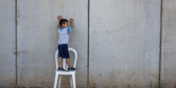 A little boy stands on a plastic chair with his hands against a wall which is the backdrop of the chair
