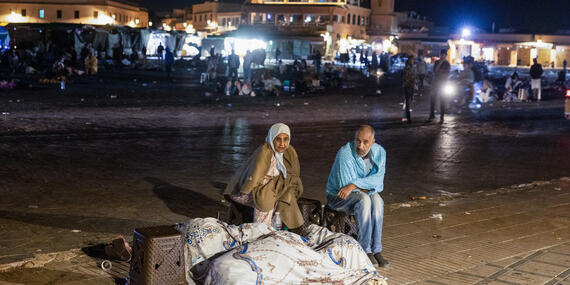 A man and a woman sit on their bags and belongings on a road.