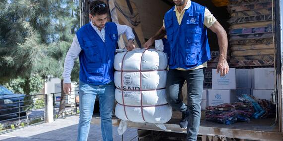 Two men in blue IOM vests unload a package labelled USAID from a truck.