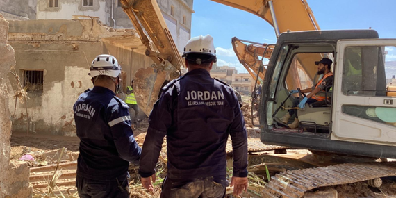 Men in safety jackets stand while another man operates a forklift amid a site of rubble and destruction.