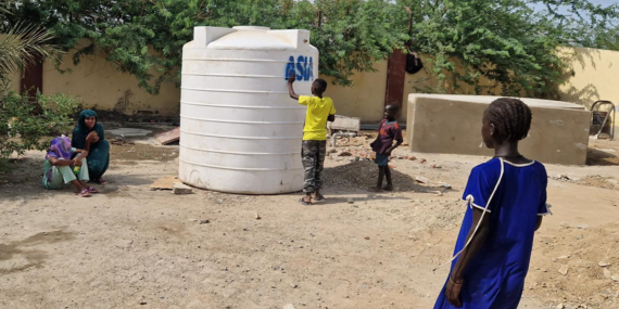 A child leans against a plastic water tank that stands on the ground.