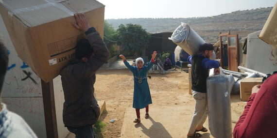 Two men carry boxes and packages, while a child looks on with her arms raised.