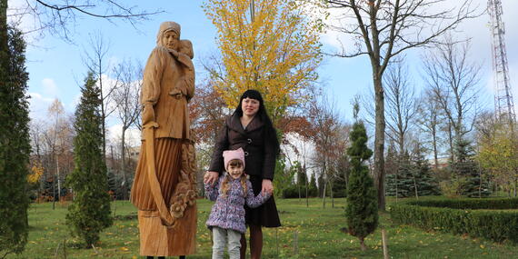 A woman with her hands around a little girl in front on her, stands in whhat appears to be a park with a statue of a mother and child in the background.