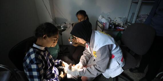 A woman in a white jacket bandages a child's leg in a dimly lit room.