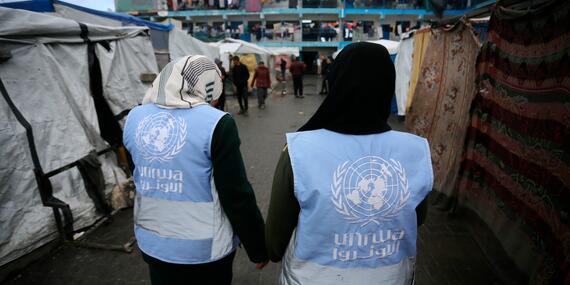 Two women wearing UNRWA vests walk down an alley. Informal shelters with people can be seen in the background.