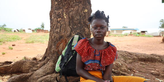 A young girl sits underneath a tree on the ground. A backpack can be seen placed behind her.