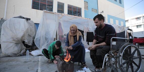A man in a wheelchair, a woman on a stool and a young boy on the ground sit near a makeshift fire in a drum in an open area outside a building. Scraps of paper lie on the ground next to the drum.