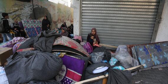 A woman with a baby on her lap sits on the ground against a shuttered building with a mound of baggage wrapped up in cloth, mattresses etc around her.. Few other women with similar bags and mattresses can be seen in the background.