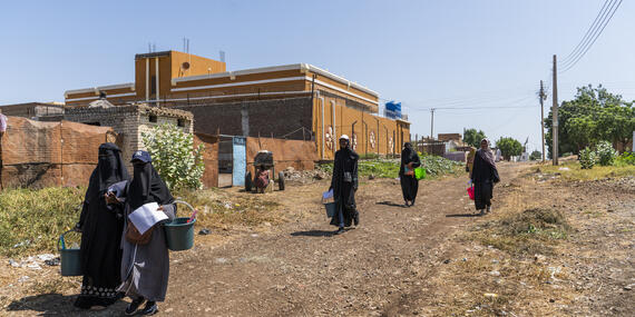 Women with plastic buckets containing packages walk down an untarred road.