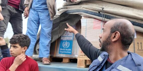 A man wearing a blue UN vest is pointing at a crate partially covered by sheeting on what appears to be the back of an open vehicle. Two men are standing on the vehicle. The exposed crate has a WHO sticker on it o