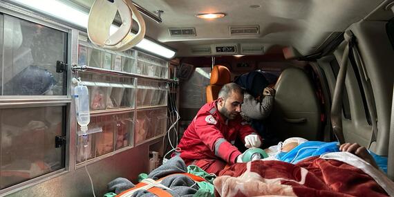 A man wearing a red jacket sits next to a stretcher with a patient inside an ambulance.