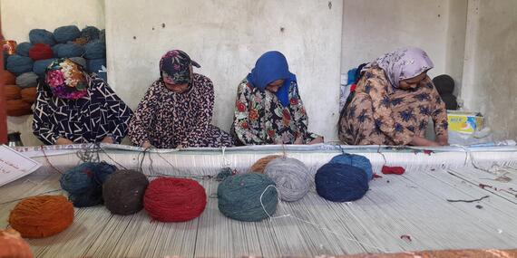 Women sit on the ground in front of a handloom placed on the ground. Big balls of wool have been placed on the loom.