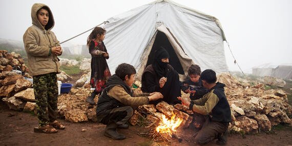 Children sit near an open fire outside a plastic covered informal tent. A heavy fog surrounds them.