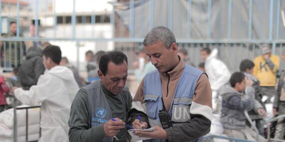 Two men - one wearing a UNRWA vest and holding a pen in his hand and the other wearing a UN vest and holding a notebook and a pen confer over the notebook in an open space. stacks of sacks in trolleys and people can be seen in the background.