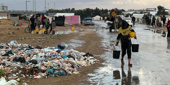 A boy walked through a flooded mud path carrying small bucket-like containers in either hand. A pile of rubbish is stacked along the path. Informal tents can be seen in the background.