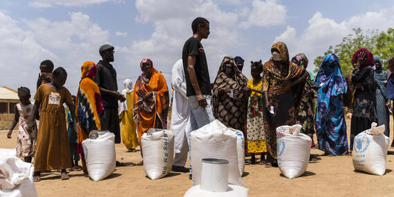 People stand in a queue under open skies on an exposed ground with sacks of grain placed next to them.