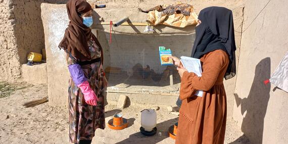 A woman holding a notebook talks to another woman in an open space outside a house.