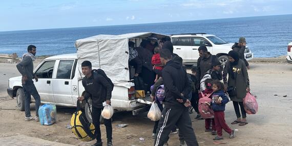men, women and children stand near a vehicle on a road along the sea. All of them hold bags. 