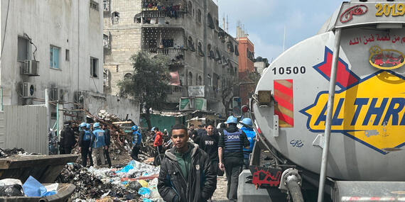A tanker is seen standing on a street littered with garbage. Damaged buildings can be seen in the background, A boy and people with blue helmets can also be seen next to the tanker.