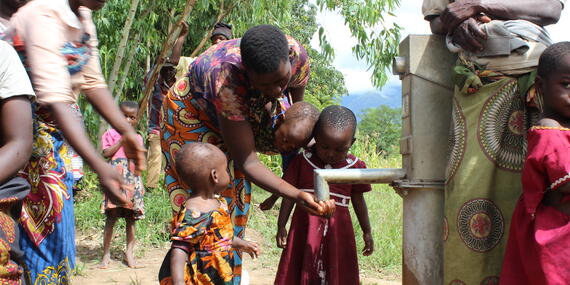 A woman helps children drink water from a tap outside.