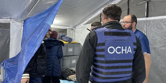 A man wearing an OCHA vest along with two mena and woman stand near a stretcher bearing a person in a tented space.