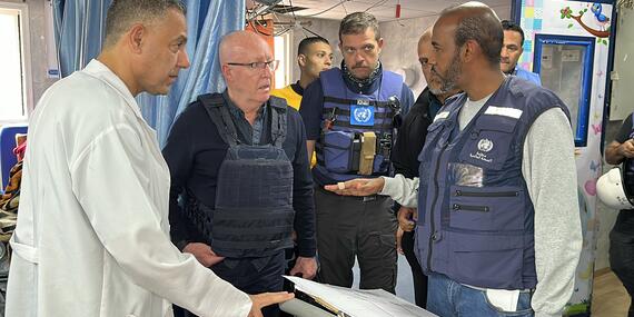 Men in blue vests stand in what appears to be the inside of a hospital. A man in a white jacket looks on.