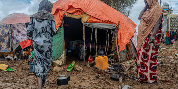 Women stand outside an informal home covered with plastic sheet. The ground is muddy and wet.