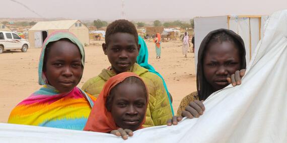 Three women and a young man look at the camera. Some of the women are clutching a plastic sheet. A UN vehicle and tents can be seen in the background.