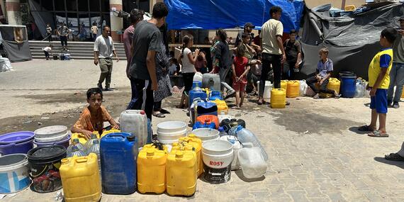 Children and adults gather around a large number of plastic containers and jugs, waiting in line to collect water outside a building in Gaza. The area is crowded, with makeshift shelters visible in the background. Gaza