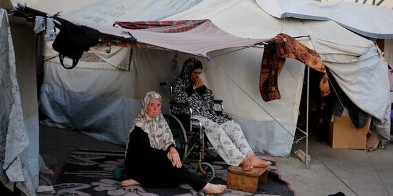 Two women sit outside their tent in a displacement camp. One woman is in a wheelchair, while the other sits on the ground. The makeshift shelter is surrounded by modest belongings and laundry, highlighting the challenging living conditions faced by displaced individuals.