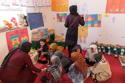 Children sit on a carpet in a classroom engaged in educational play activities with a woman watching over them, while another woman is putting something up on a wall, which is covered with colourful educational posters.