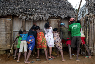 Men, women and children help prop up the wall of a house made up of grass and wood.