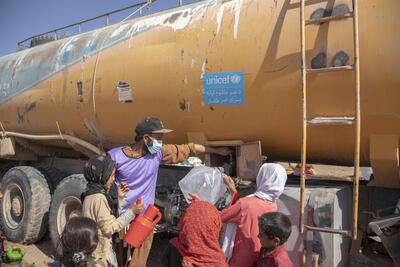 A man stands near a tanker with a UNICEF sticker on it as women and children fill water.
