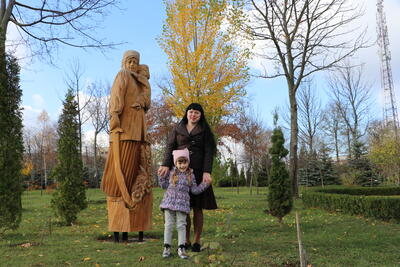 A woman with her hands around a little girl in front on her, stands in whhat appears to be a park with a statue of a mother and child in the background.