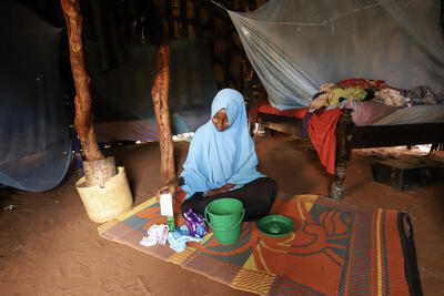 A girl seated on a mat with a plastic bucket and a packet of sanitary napkins, women's underwear, a bottle of coconut oil and a comb laid out on the mat.