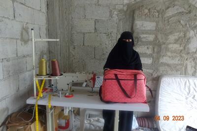 A woman stands behind a worktable. A leather bag , a sewing machine and big spools of thread are on the table.