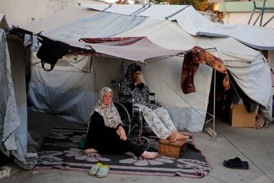 Two women sit outside their tent in a displacement camp. One woman is in a wheelchair, while the other sits on the ground. The makeshift shelter is surrounded by modest belongings and laundry, highlighting the challenging living conditions faced by displaced individuals.