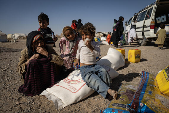 Women and children sit on an open ground with sacks of food.