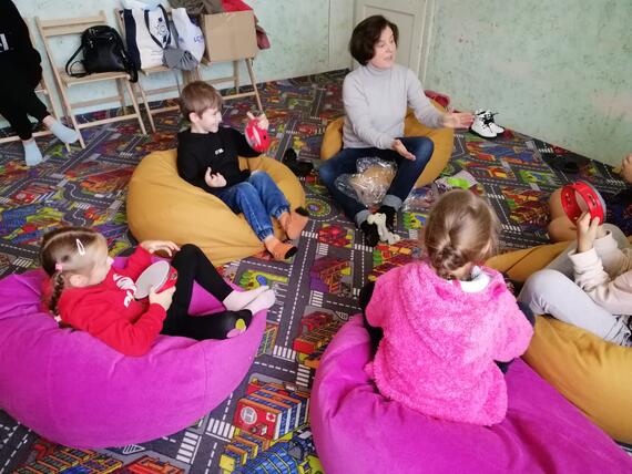 A woman seated on a floor cushion is interacting with four children also seated on floor cushions in a carpeted room.