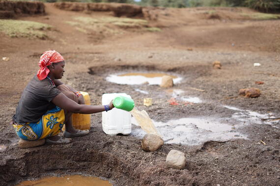 A woman is filling water in a container. She is sitting near a small pool of muddy water.