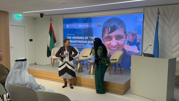 Two women stand on a raised platform. One is dancing and the other  is holding a microphone in her hand.  A digital banner behind them reads the opening of the humanitarian hub