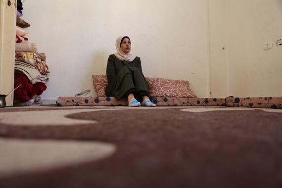 A woman sits on a mattress on a carpeted floor.