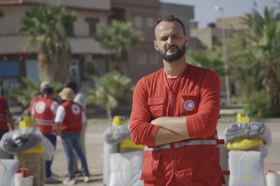 A man in red overalls with a Red Crescent faces the camera. People in red vests can be seen in the background.