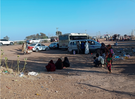 Women seated on the ground in an open space. People can be seen getting into vehicles in the background.