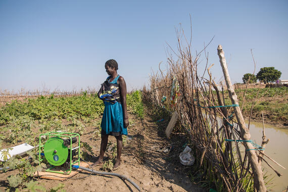 A woman stands in a field with plants. A pump with a pipe is near her feet.