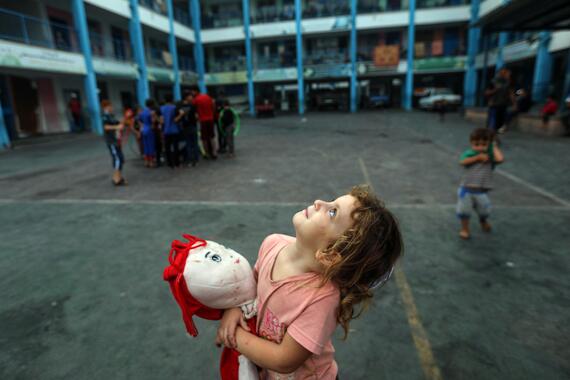 A little girl stands in a compound of a building holding a stuffed toy and looks up 
