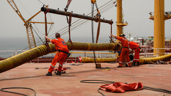 Men in orange overalls are working on huge pipes standing on the deck of a ship. Sea can be seen in the background.