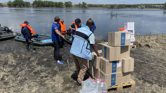 Men with life jackets stand next to a canoe near a body of water, while a man in blue UN vest stands next to a pile of boxes