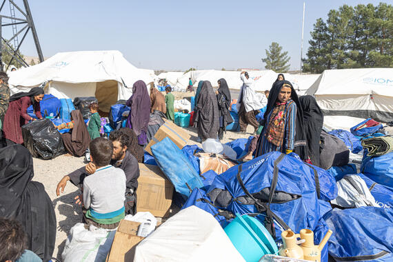 People stand with packages next to them in an open area with tents.